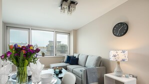 A modern living room with a sofa, a dining table set for two, a vase of flowers, and a wall clock. - Gorgeous 5 Star Location 2 min to Brighton Beach (Brighton)