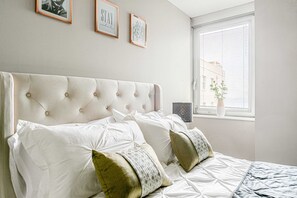 A bedroom with a tufted headboard, white bedding, and a window with blinds.