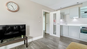 A modern living room with a flat-screen TV on a wooden stand, a small potted plant, and a wall clock. A kitchen with white cabinets and stainless steel appliances is visible through an open doorway. - The Lanes Stellar Stay Sea View 2min Walk to Beach (Brighton)