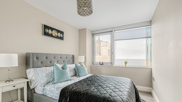A bedroom with a grey upholstered bed, a white nightstand, a black quilted bedspread, and a window with blinds. - The Lanes Stellar Stay Sea View 2min Walk to Beach (Brighton)