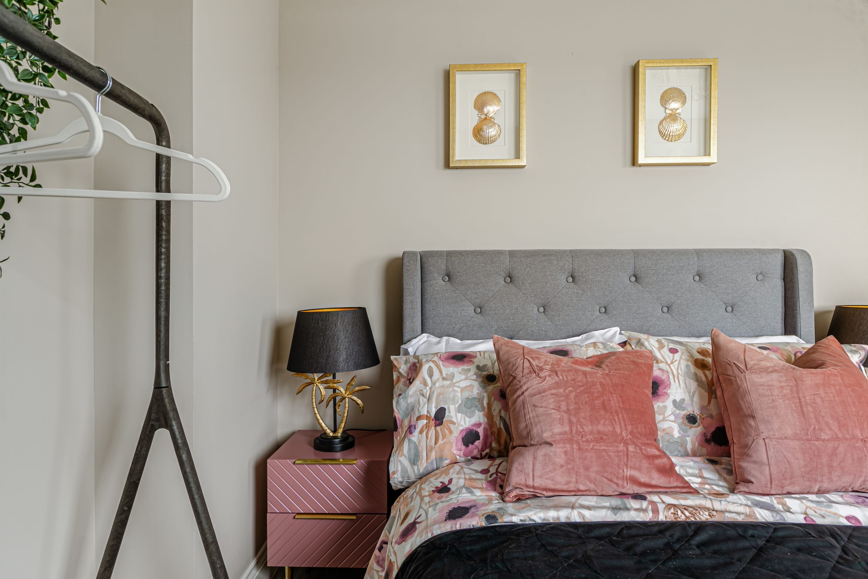 Um quarto bem arrumado com cabeceira estofada cinza, colcha com padrão floral e duas fotos emolduradas na parede.