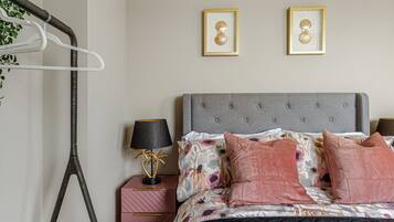 Um quarto bem arrumado com cabeceira estofada cinza, colcha com padrão floral e duas fotos emolduradas na parede.