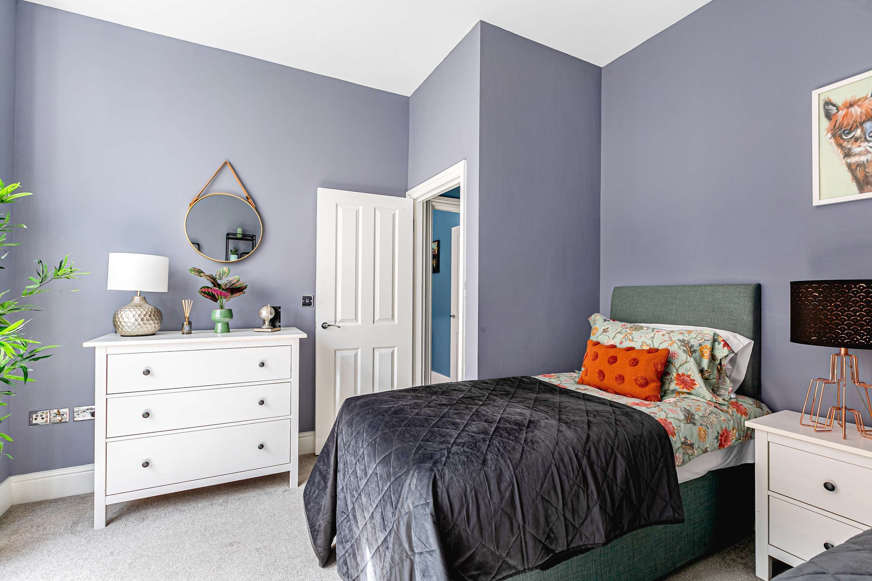 A bedroom with a bed, bedside table, dresser, mirror, and a door leading to another room.