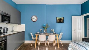 A compact kitchen with modern appliances, a dining area with a table and chairs, a clock, and a framed picture on the wall. - The Lanes Comfy Condo Walk to Beach King Bed (Brighton)
