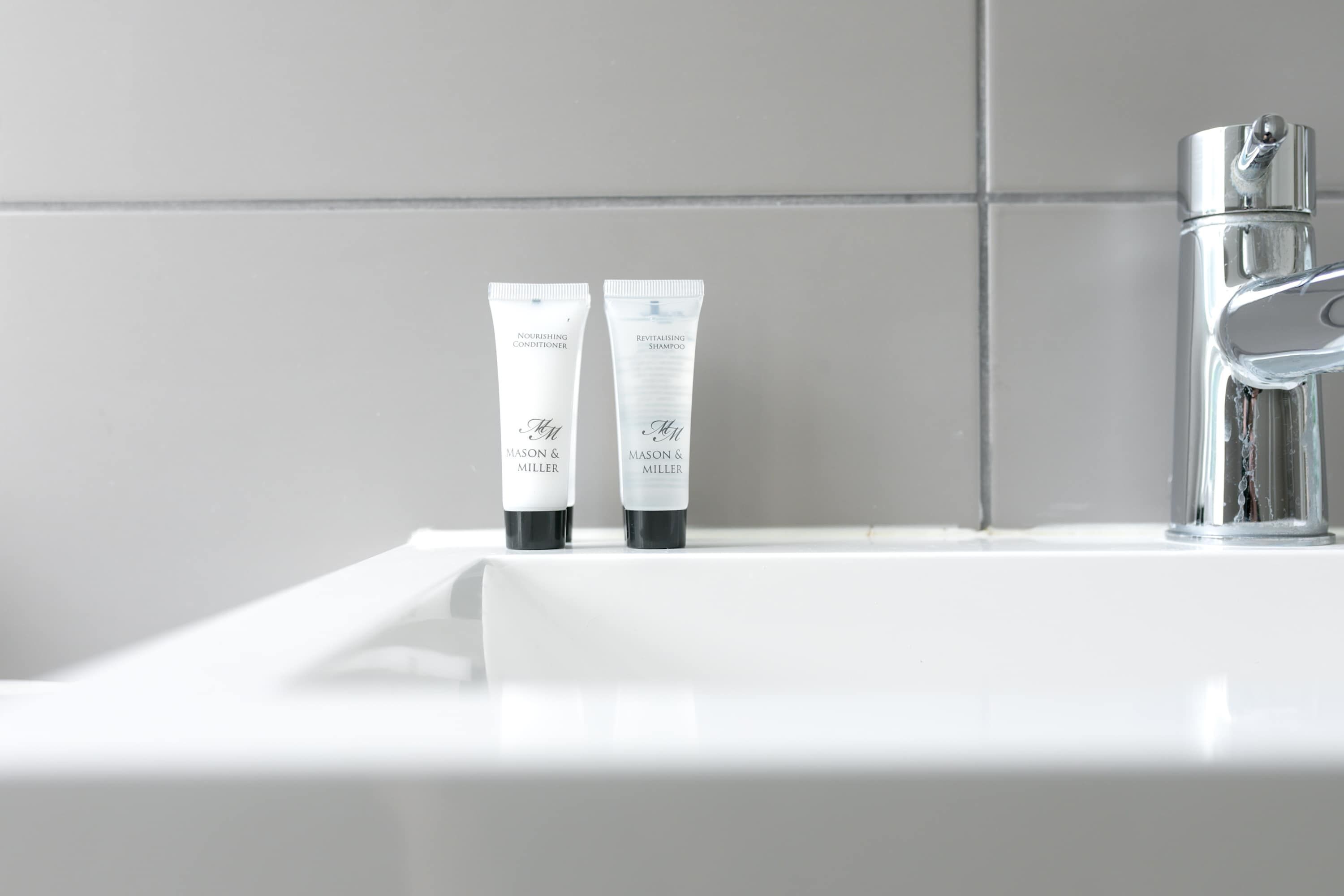 A bathroom sink with two toiletry bottles labeled 'NOURISHING CONDITIONER' and 'REVITALISING SHAMPOO' by Mason & Miller.