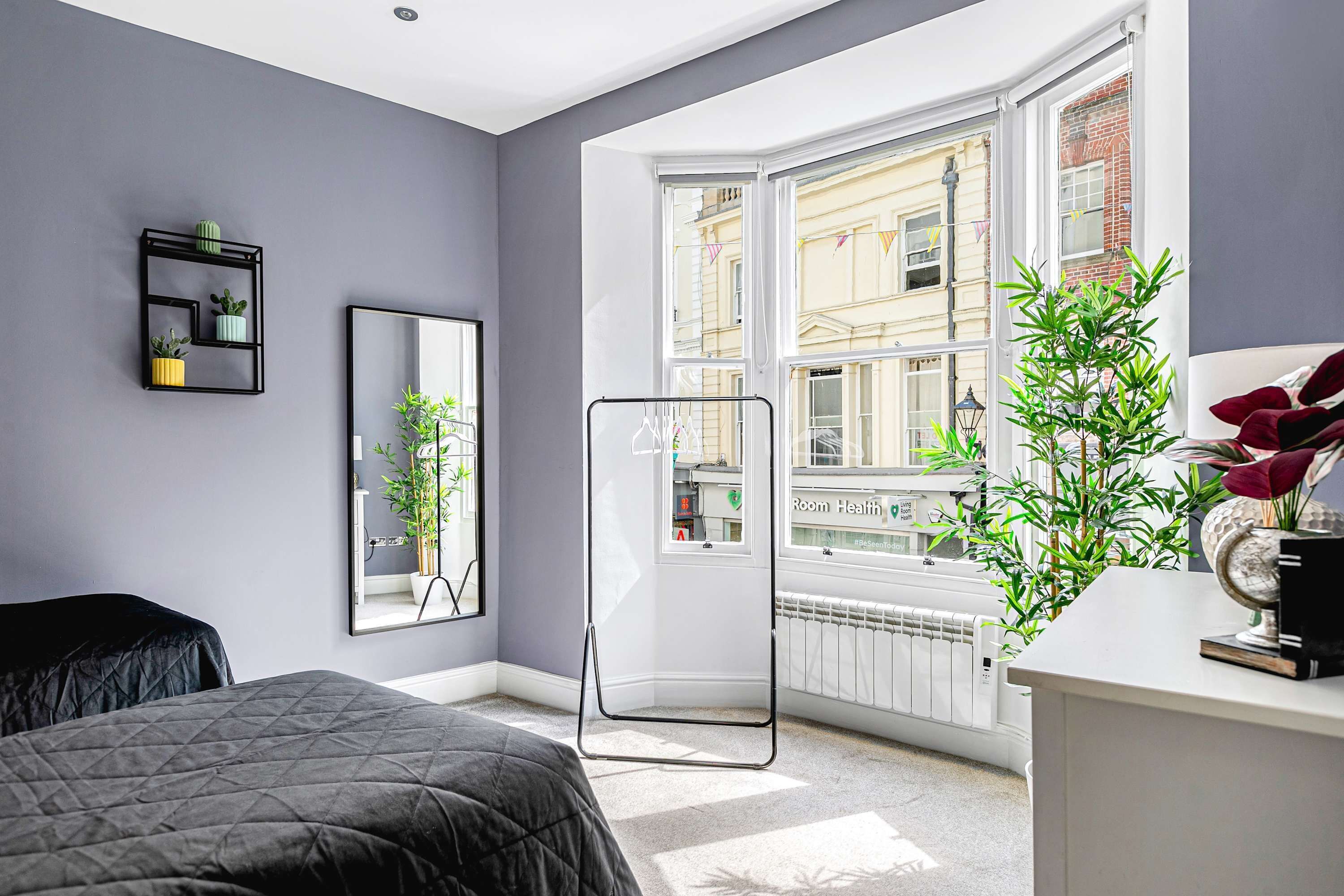 Un dormitorio moderno con un ventanal grande, una cama, una mesita de noche, un espejo y una planta.