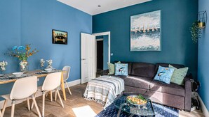 A room with a blue wall, a sofa with cushions, a dining table with chairs, and a painting above the sofa. - The Lanes Comfy Condo Walk to Beach King Bed (Brighton)