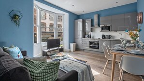 A modern kitchen with grey cabinets, a built-in oven, and a microwave. There is a dining area with a glass table and chairs. - The Lanes Comfy Condo Walk to Beach King Bed (Brighton)