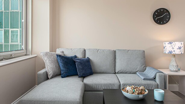 Un salon moderne avec un canapé d’angle gris, une petite table d’appoint, une horloge murale et une fenêtre avec des stores.