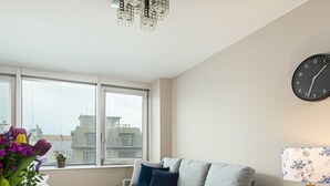 Un salon moderne avec un canapé gris, une table à manger dressée avec de l’argenterie, un vase de fleurs et une vue sur la ville à travers de grandes fenêtres.