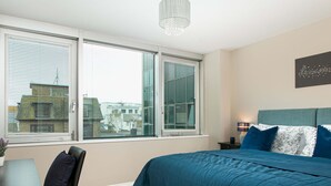 Une chambre avec un grand lit, un bureau et une chaise. Il y a deux fenêtres avec des stores, un lustre et une œuvre d’art murale.