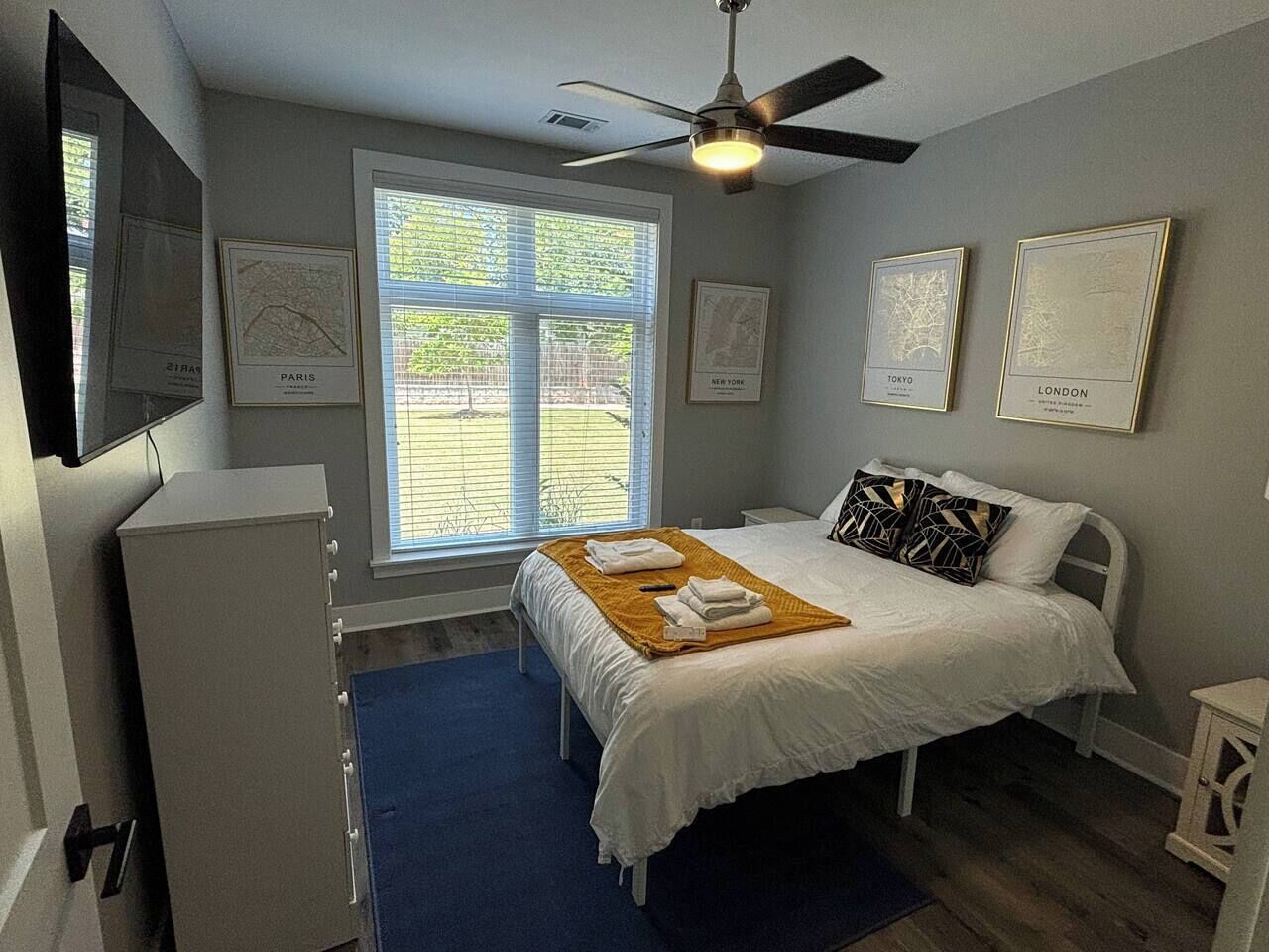 A bedroom with a bed, a ceiling fan, a window with blinds, and framed maps on the wall.