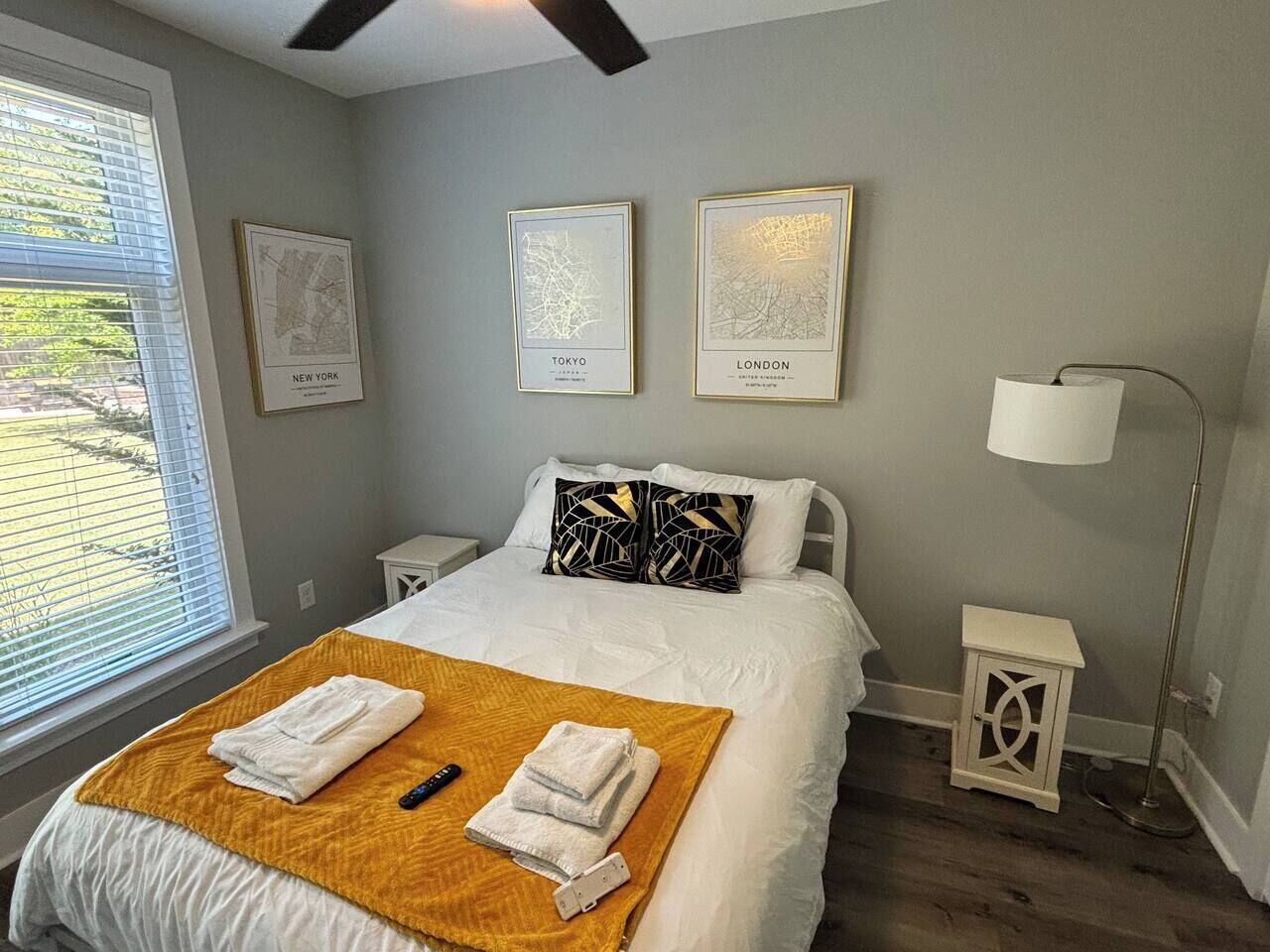 Um quarto com cama, janela com persianas, mesa de cabeceira, abajur e três mapas emoldurados na parede.