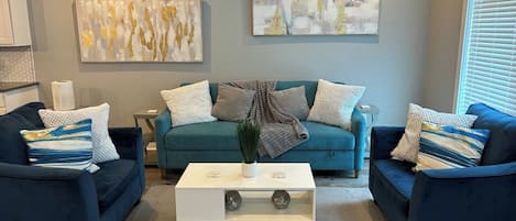 A living room with a teal sofa, two armchairs, a white coffee table, and abstract wall art.