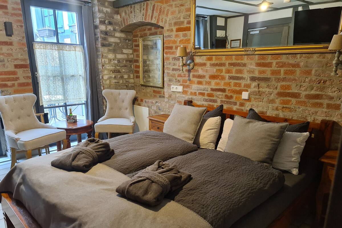 A bedroom with a brick wall, a large bed with multiple pillows, two chairs, a small table, and a mirror.