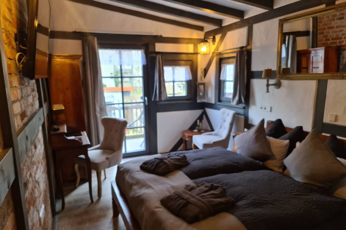 A cozy room with two beds, a desk, and a chair. There are windows with curtains, a lamp, and a mirror on the wall.