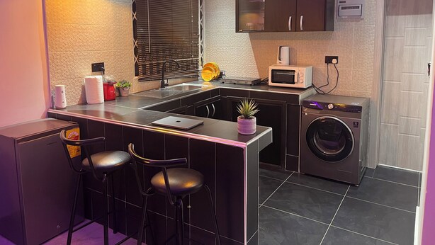 Cocina compacta con lavadora integrada, microondas y lavamanos. Hay dos taburetes de bar y una pequeña planta en maceta sobre la encimera.