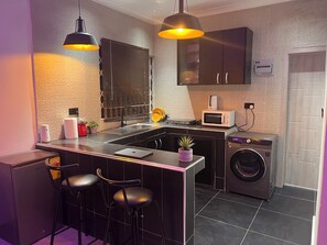 Cocina compacta con lavadora integrada, microondas y lavamanos. Hay dos taburetes de bar y una pequeña planta en maceta sobre la encimera.