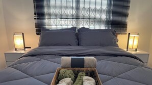 Una cama bien tendida con un edredón gris, dos lámparas de noche y una cesta de mimbre con toallas y una manta sobre una mesita blanca.