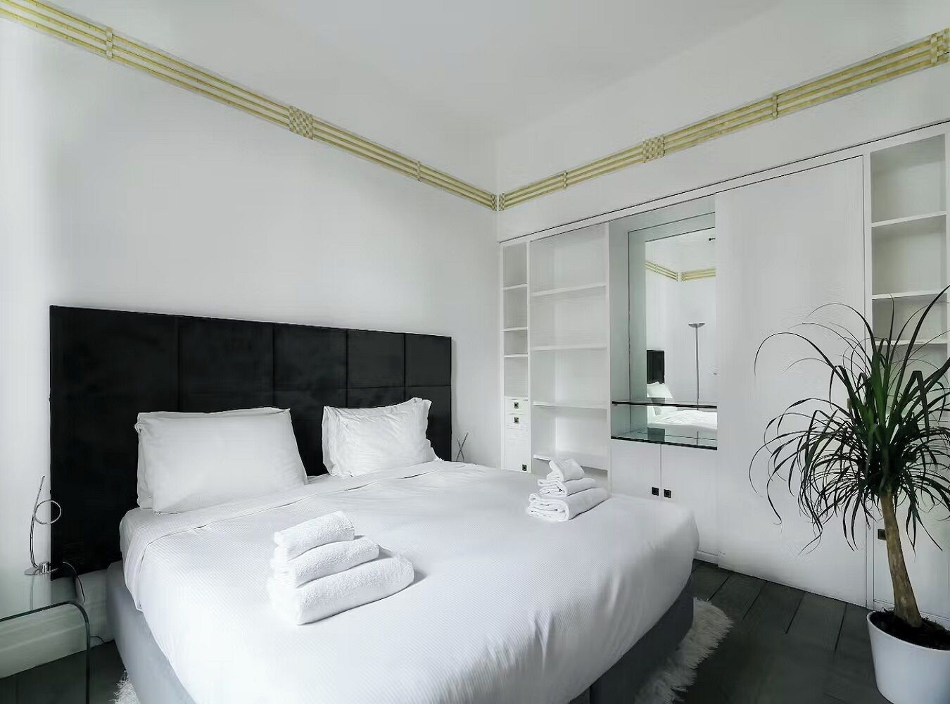 A modern bedroom with a large bed, white linens, a dark headboard, a bedside table with a lamp, a mirror, and a potted plant.