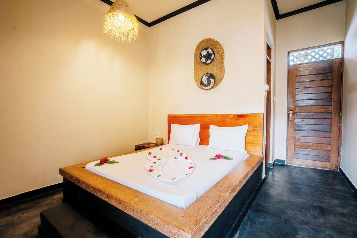 Una cama impecablemente arreglada con sábanas blancas y un plato decorativo, cabecera de madera, lámpara de fibra y puerta de madera con diseño de celosía.