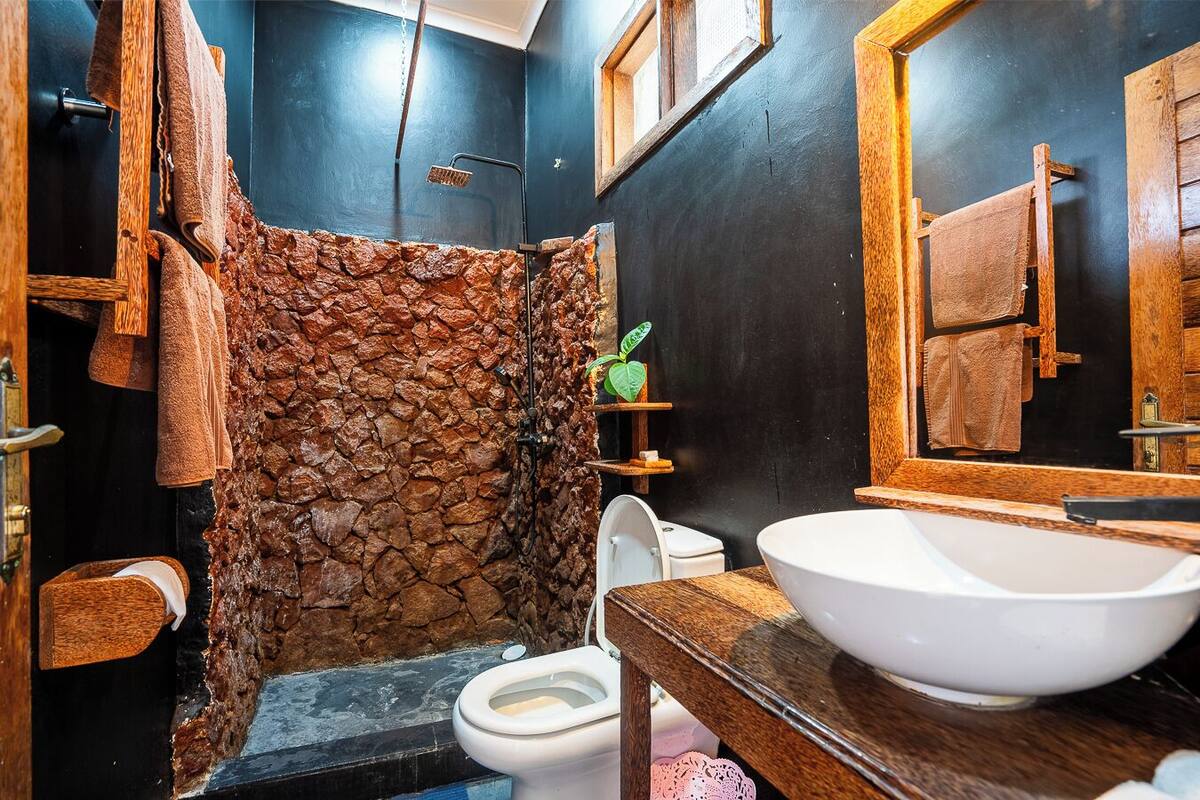 Un baño con cabina de ducha de piedra, inodoro blanco y tocador de madera con un lavabo blanco grande.