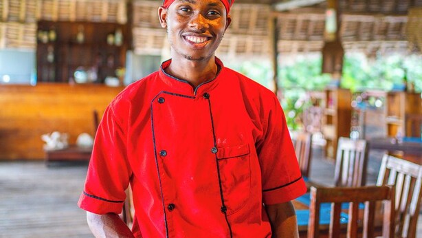 A person in a red chef's uniform standing in a restaurant with a thatched roof and wooden furniture. - La Fotana Restaurant And Bungalows (Kendwa)