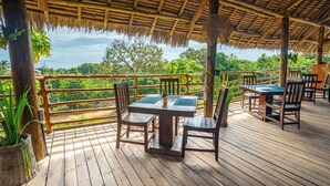 An outdoor wooden deck with thatched roof seating and tables, overlooking lush greenery. - La Fotana Restaurant And Bungalows (Kendwa)
