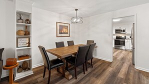 Ein Esszimmer mit Holztisch und sechs Stühlen, eine weiße Regalanlage mit Dekoration und eine durch einen offenen Durchgang sichtbare Küche.