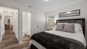Ein Schlafzimmer mit einem großen Bett, einem Nachttisch mit Lampe, einem Fenster mit Jalousien und einer Tür zu einem anderen Raum.