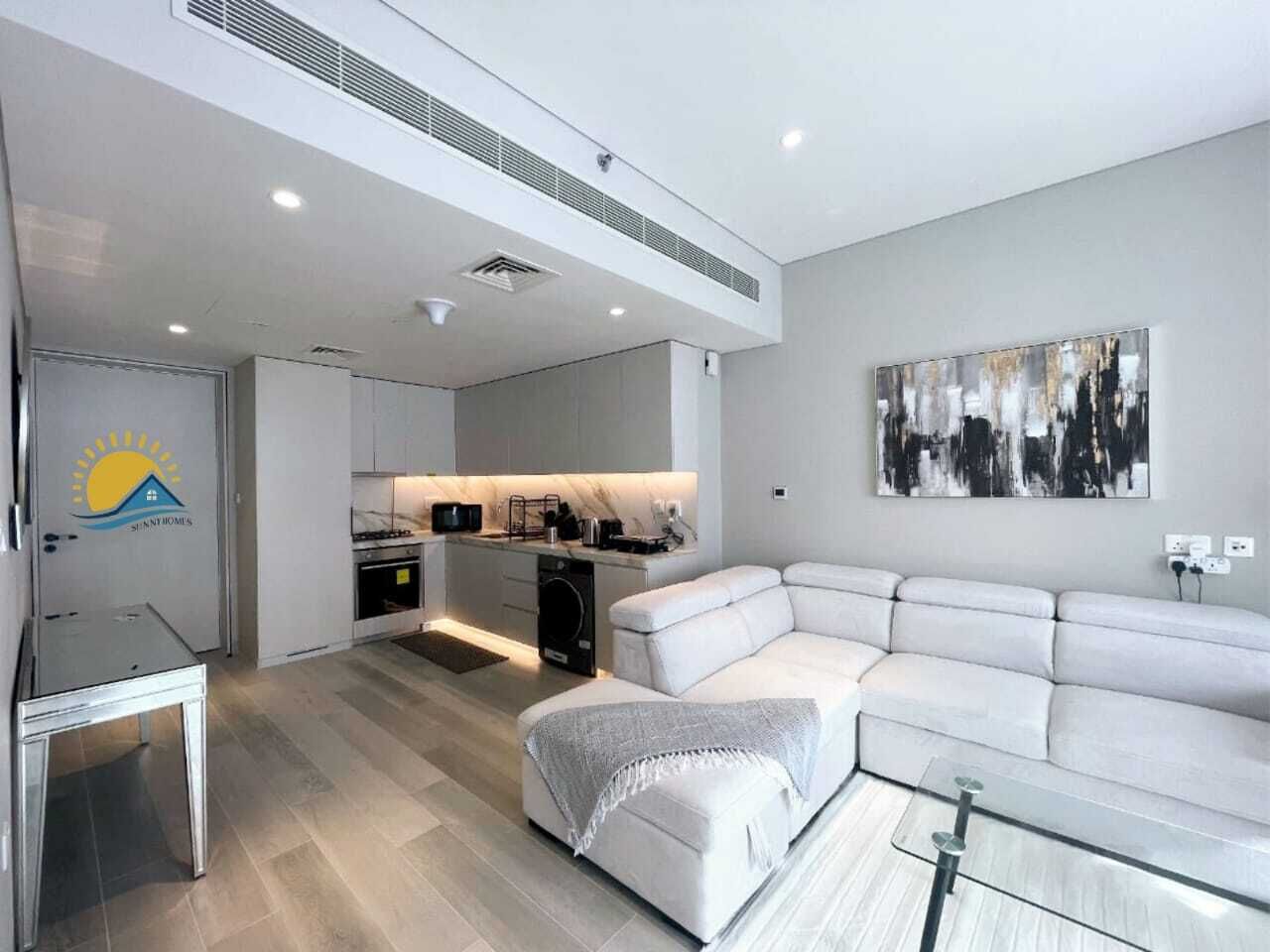 Ein modernes Wohnzimmer mit einem weißen Sofa, einem Glastisch und einer Küchenzeile mit Mikrowelle und Herd.