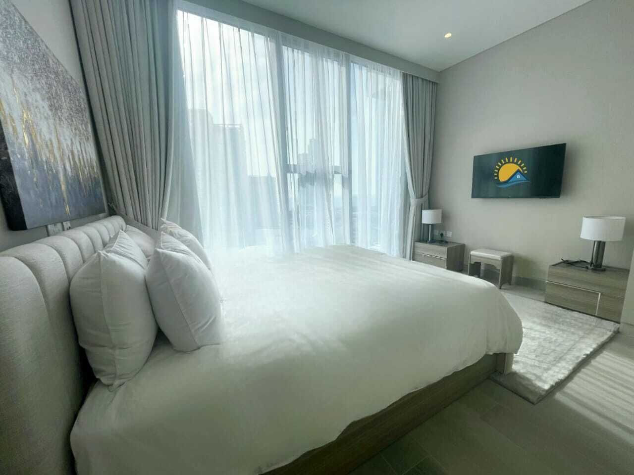 A modern hotel room with a large bed, a flat-screen TV, and a view of the city through sheer curtains.