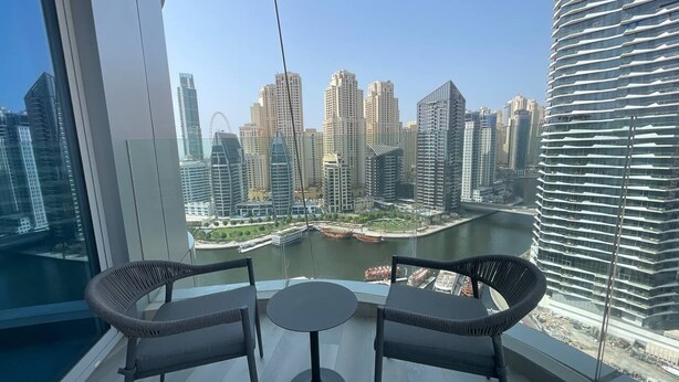 A balcony with two wicker chairs and a small round table, overlooking a cityscape with modern buildings and a river.