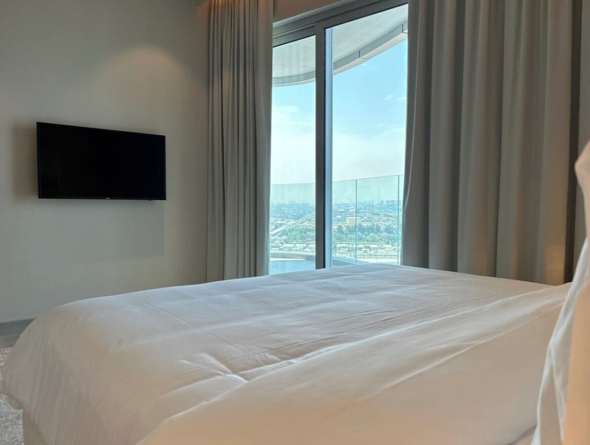 A hotel room with a large bed, a flat-screen TV mounted on the wall, and a view of a cityscape through a large window.