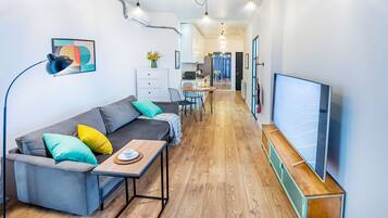 Uma sala moderna com sofá, mesa de centro e televisão. Cozinha visível ao fundo, com área de refeições.
