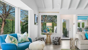 A spacious living room with a blue sofa, a white furry ottoman, and a beige sofa. There are two round tables with zebra-patterned stools. The room has large windows and a view of greenery outside. - The Colony Hideaway - Pool Wifi Spa Private (Palm Springs)