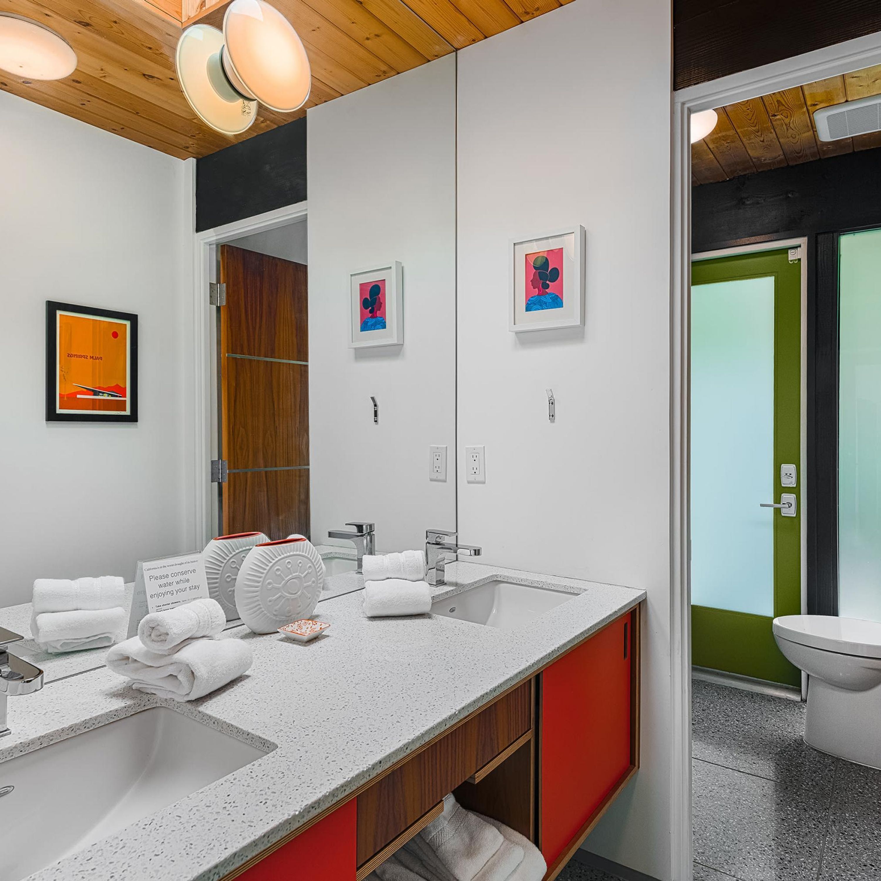 A modern bathroom with a large mirror, two sinks, and a toilet. There are towels and a sign about water conservation.