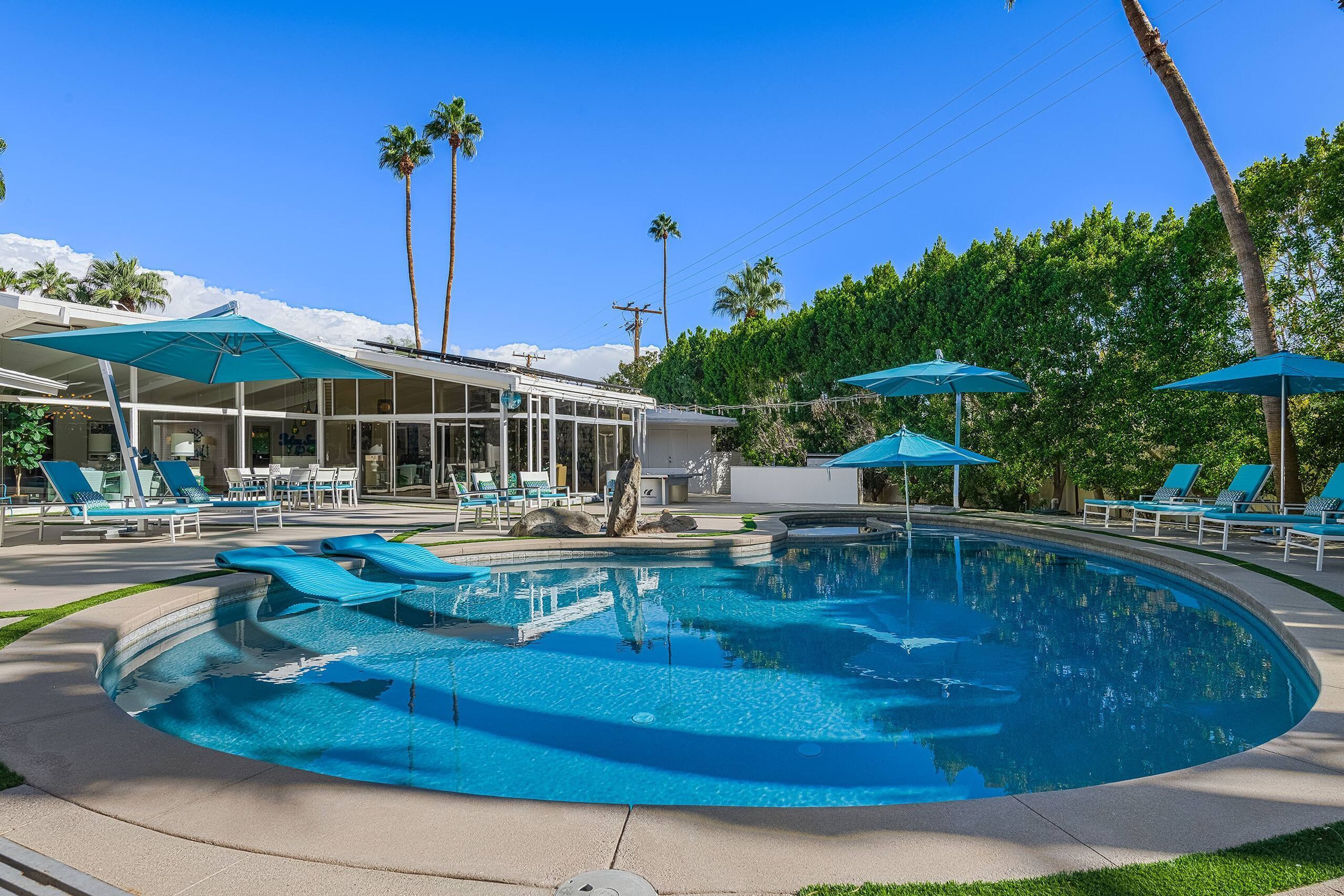 A swimming pool with lounge chairs and umbrellas, surrounded by palm trees and a building with a glass facade.