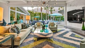 Una sala de estar moderna con sofá, mesa de centro y sillones, un televisor grande y vistas a una piscina y montañas.