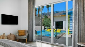 Un soggiorno moderno con una grande televisione a schermo piatto, un'area salotto accogliente con divano e poltrona, e vista su piscina e montagne attraverso ampie vetrate a tutta altezza.