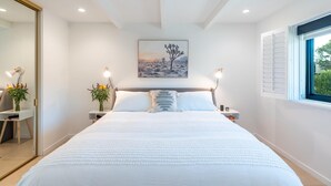 Une chambre avec un grand lit, deux tables de chevet avec des lampes, un tableau au mur et une fenêtre avec des volets.