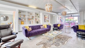 Ein modernes Wohnzimmer mit einem violetten Sofa, schwarzen Ledersesseln und einem Glastisch. Weiße Regale mit Dekorationen und ein großes Fenster mit Blick ins Freie.