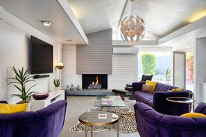 A modern living room with a fireplace, purple sofas, a glass coffee table, and a decorative chandelier. - The Yellow Door on Shadow Mountain (Palm Desert)