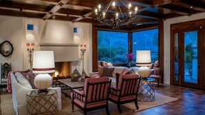 A cozy living room with a fireplace, comfortable seating, and a large window offering a view of the outdoors. - Villa Vista - Spanish Luxury Pool Wifi (Palm Springs)