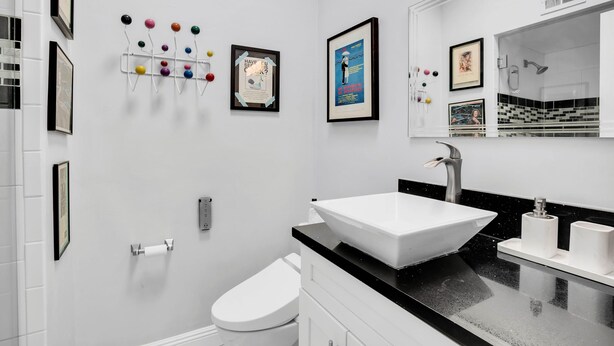 A modern bathroom with a white sink, black countertop, and a large mirror. There are framed pictures on the wall and a toilet paper holder. - Escapade in Palm Springs (Palm Springs)