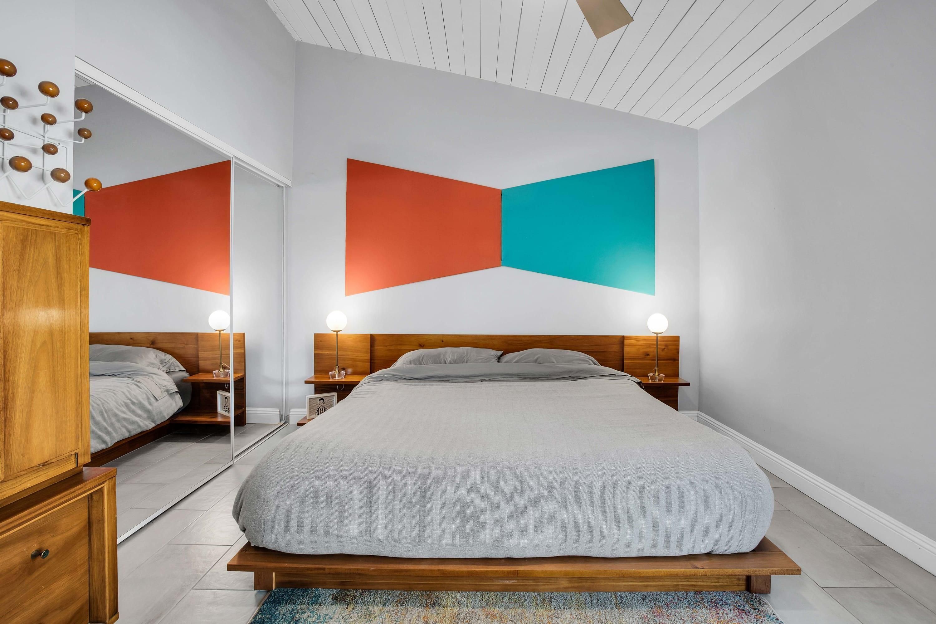 A modern bedroom with a large bed, wooden headboard, bedside tables with lamps, and a colorful abstract wall art piece.