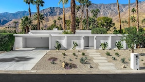 Ein modernes Gebäude mit heller Fassade, ein angelegter Garten mit verschiedenen Pflanzen und eine Bergkulisse im Hintergrund.