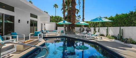 A modern pool area with lounge chairs and umbrellas, surrounded by palm trees and a white building.