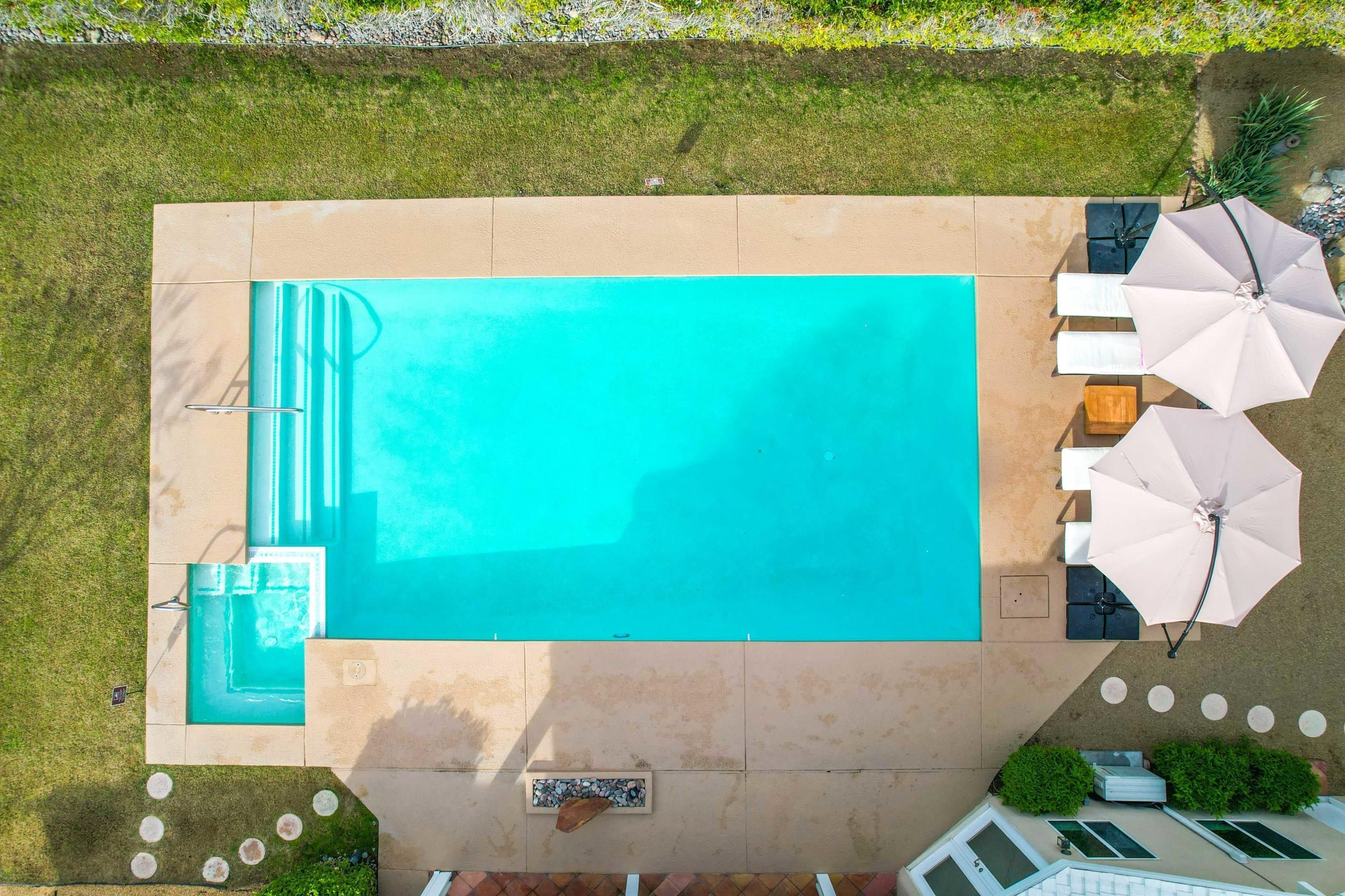 A rectangular swimming pool with a tiled deck, surrounded by a grassy area and a patio with outdoor furniture and umbrellas.