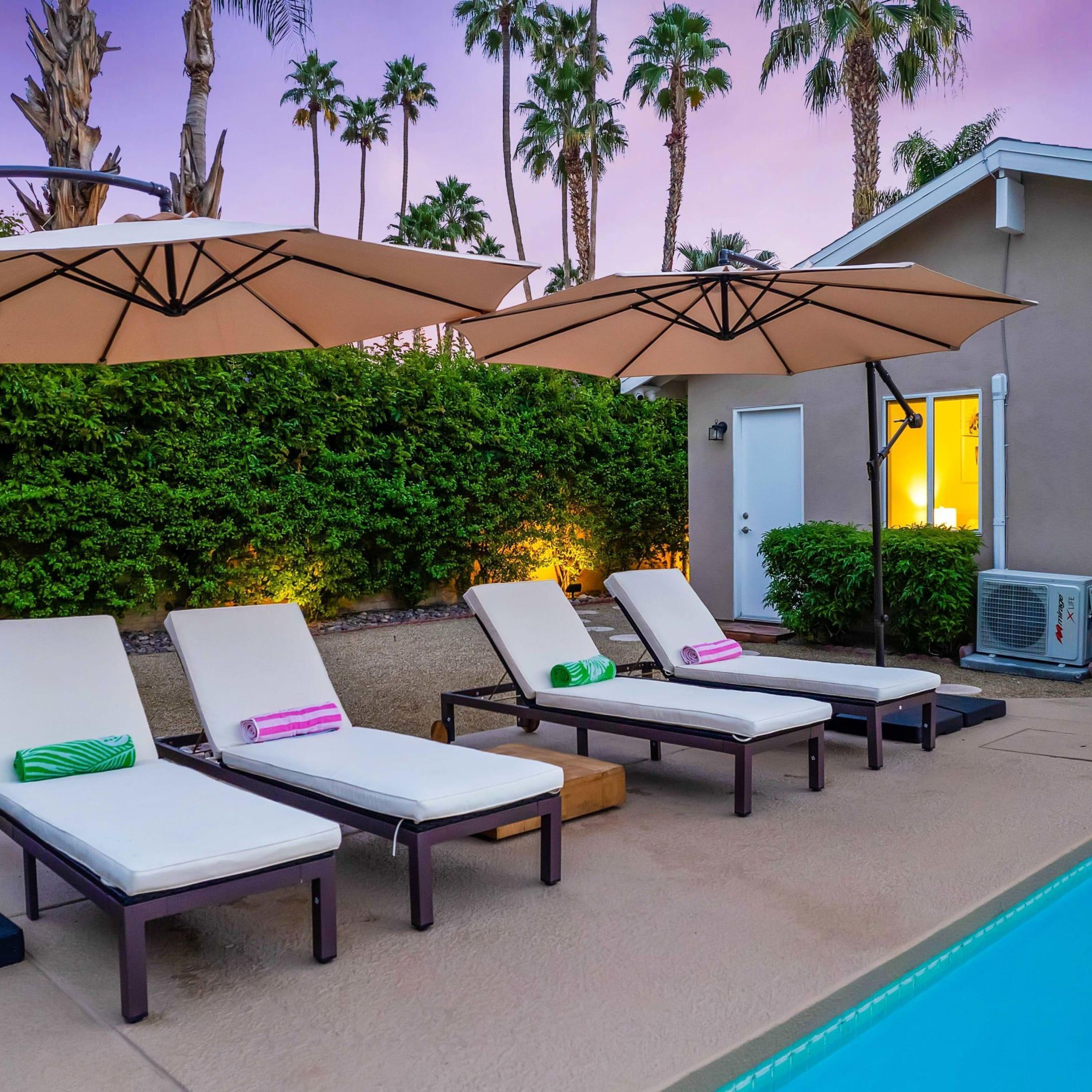 A poolside area with lounge chairs and umbrellas, a hedge, and a building in the background.